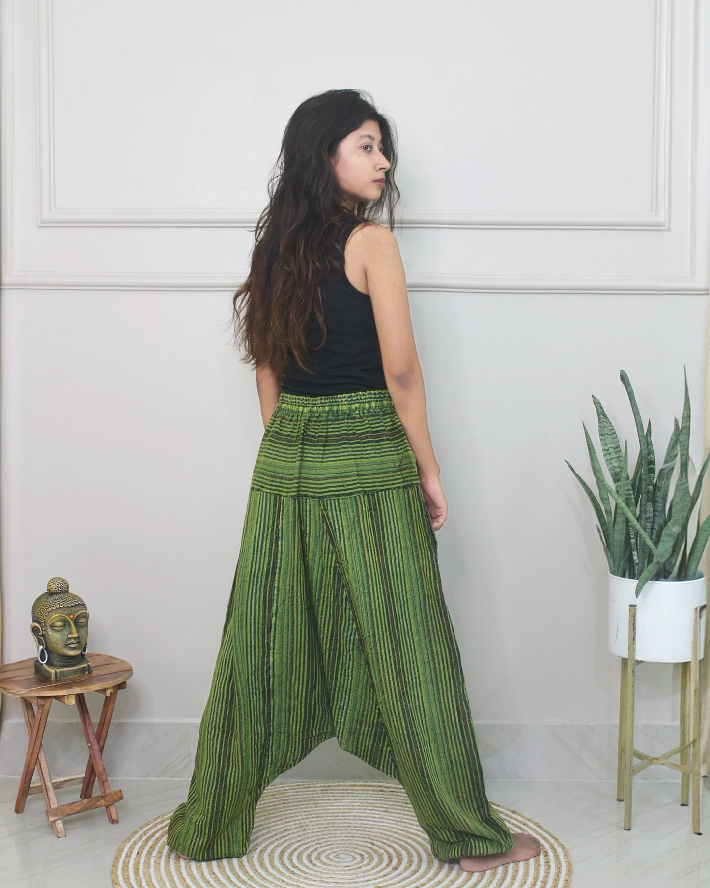 Woman wearing green striped harem pants and a black top in a room with a white wall and small table.