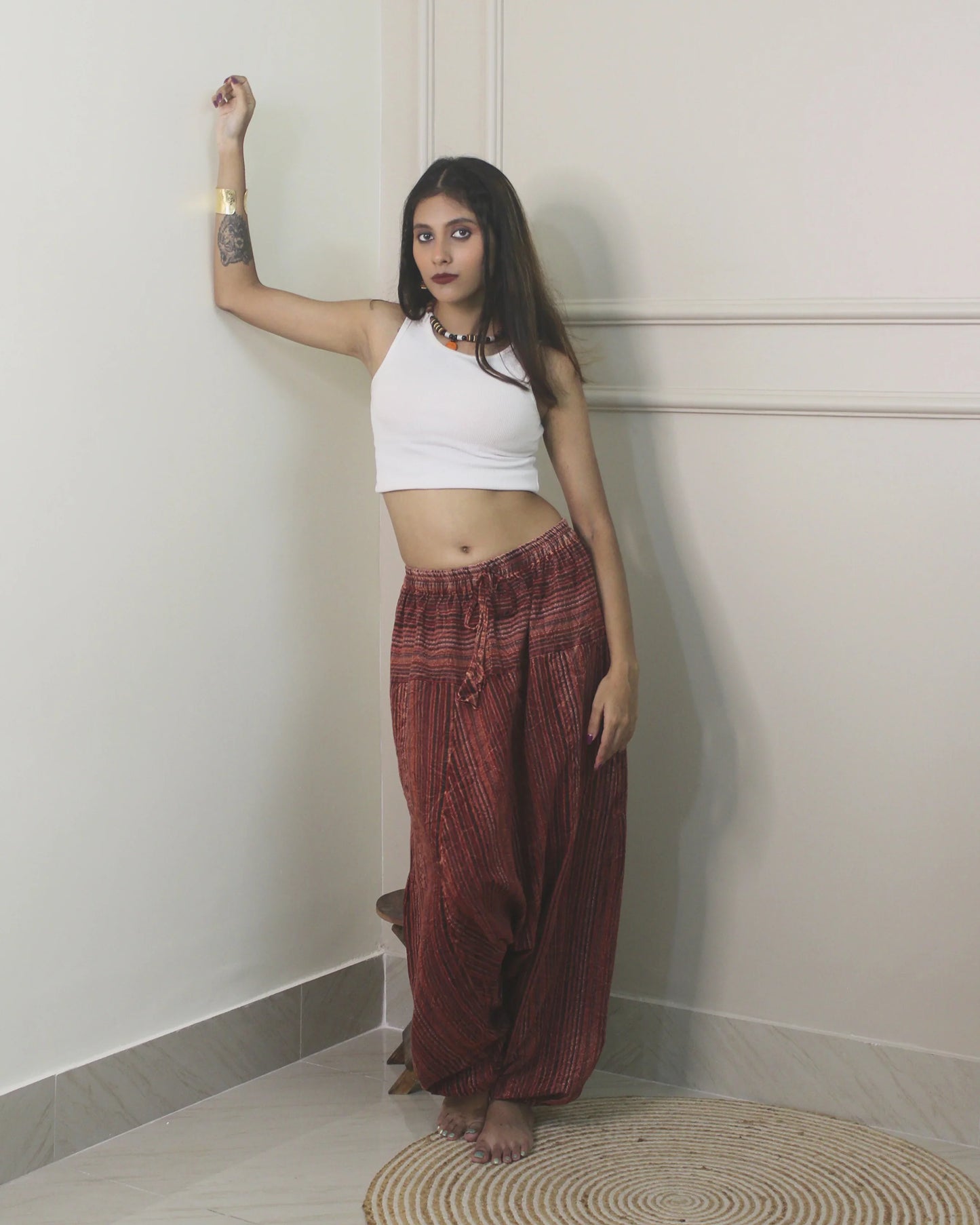 Woman wearing a white top and harem pants standing against a plain wall.