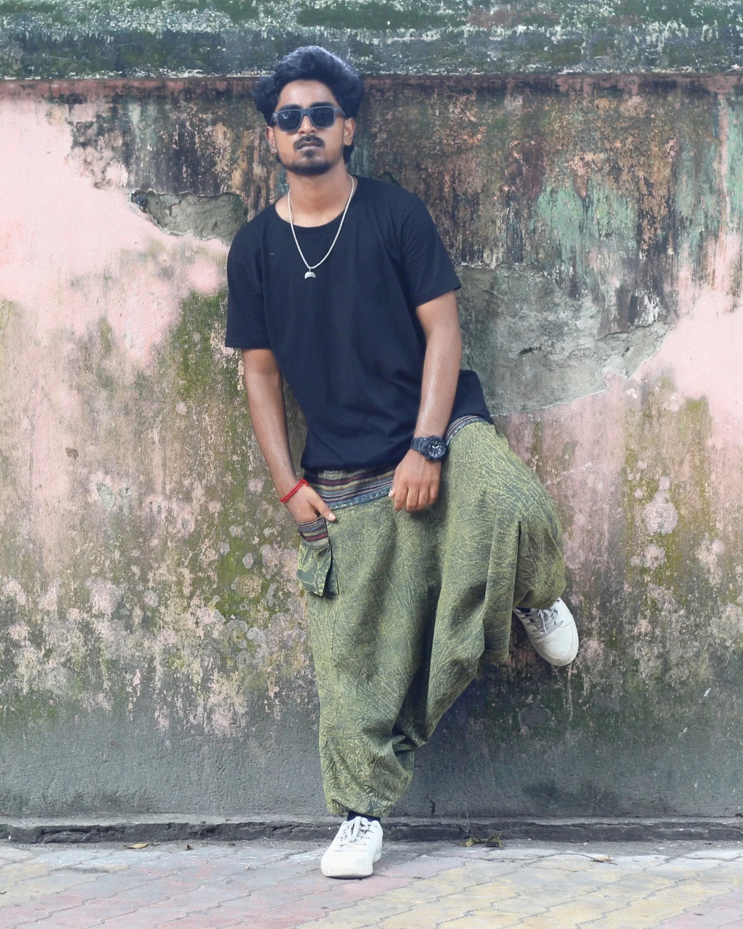 Man wearing sunglasses, black shirt, green harem pants, and white sneakers against a textured wall.