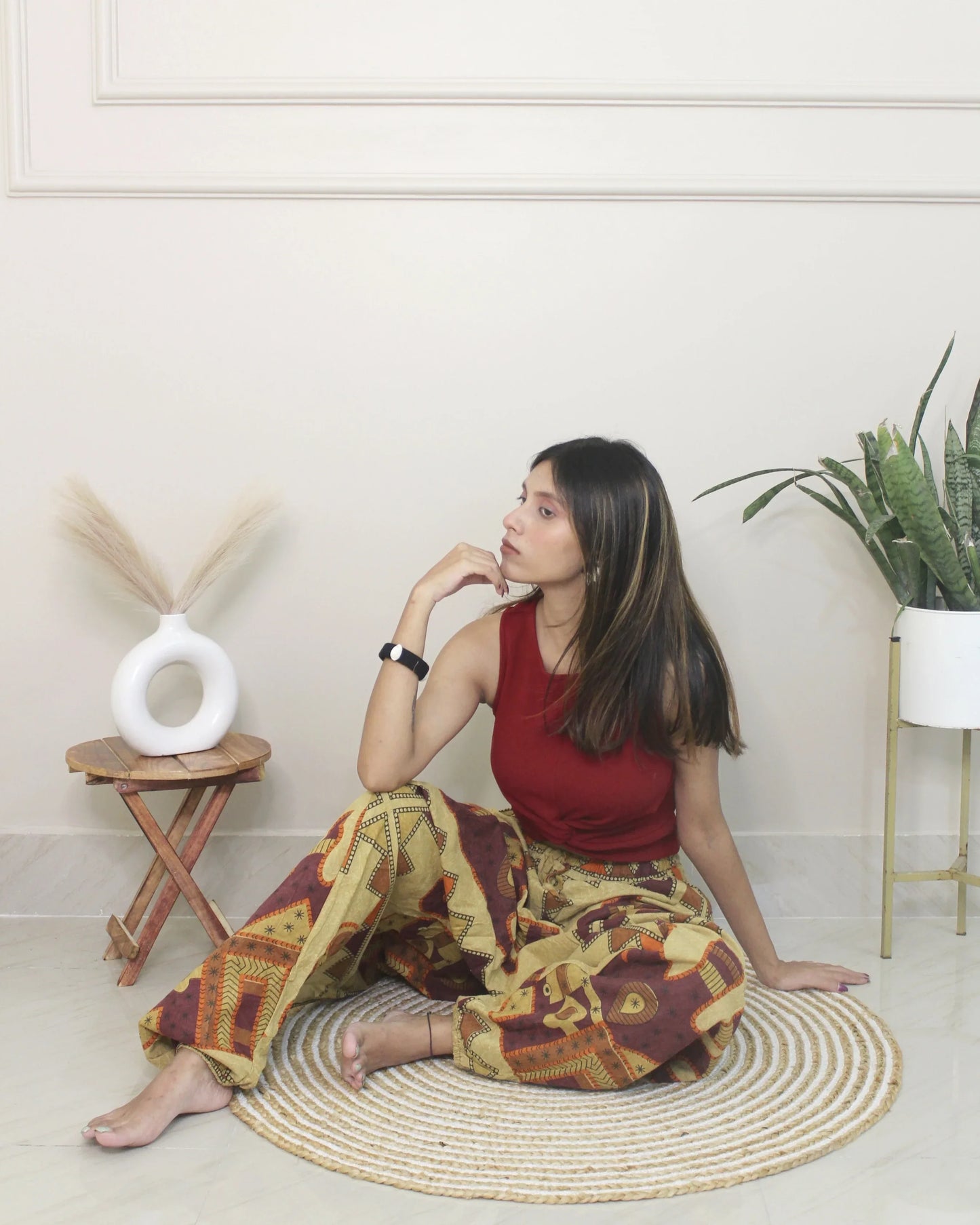 Woman sitting wearing a harem pants on a woven mat in a room with plants and a small table.