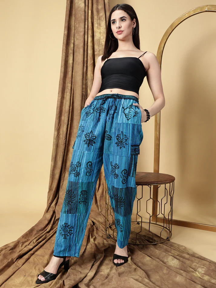 Woman wearing a black top and blue patterned pants standing against a beige curtain.