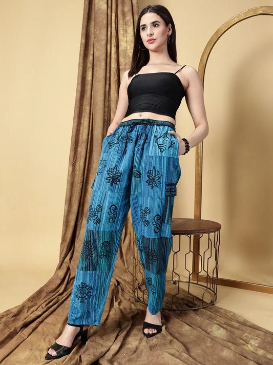 Woman wearing a black top and blue patterned pants standing against a beige curtain.