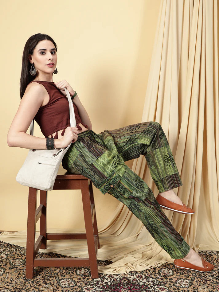 Woman sitting on a stool wearing a brown top and green patterned pants, holding a white bag.