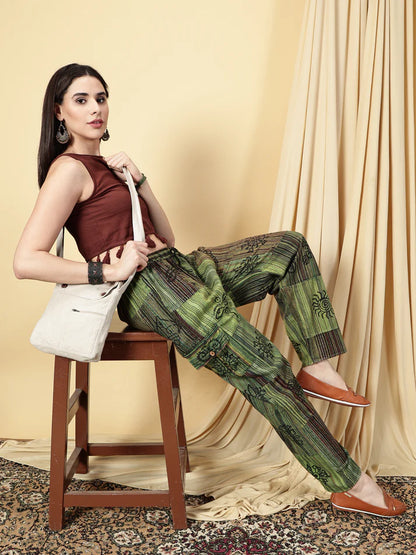 Woman sitting on a stool wearing a brown top and green patterned pants, holding a white bag.