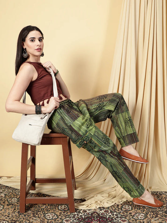 Woman sitting on a stool wearing a brown top and green patterned pants, holding a white bag.