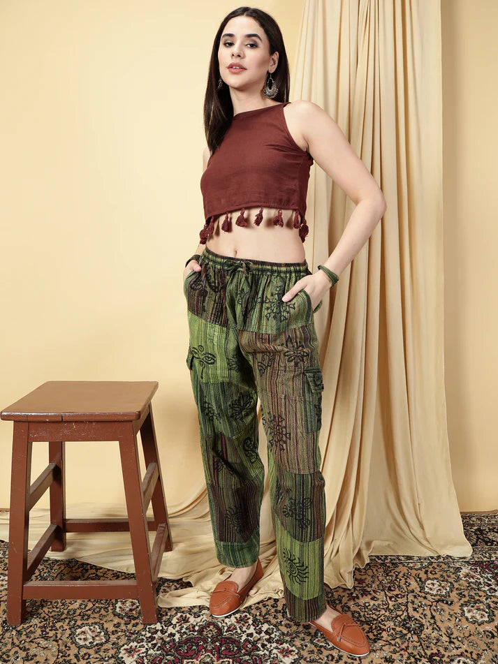 Woman wearing a brown top and green patterned pants standing in a room with a wooden stool and beige curtain.