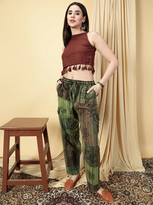 Woman wearing a brown top and green patterned pants standing in a room with a wooden stool and beige curtain.