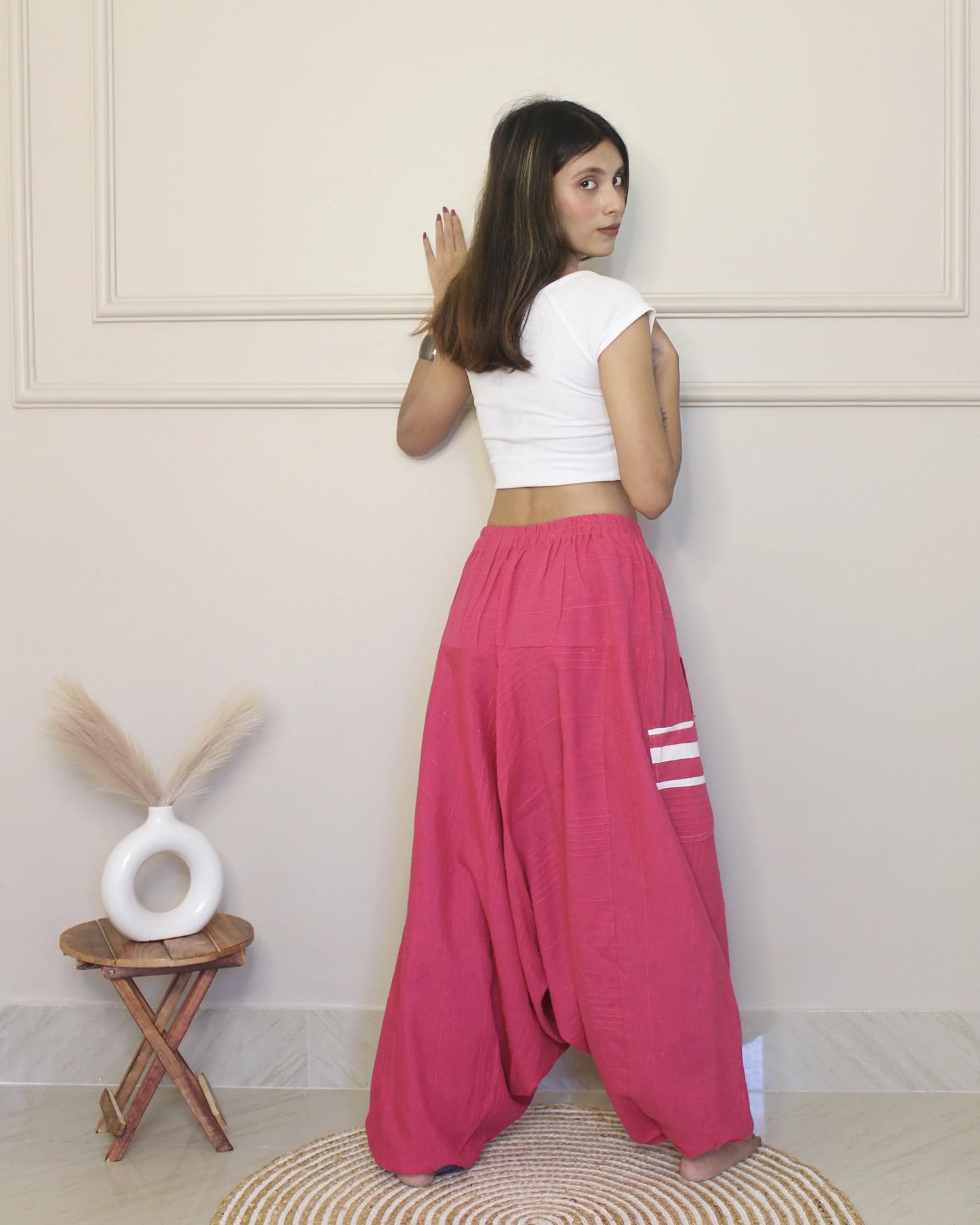 Woman wearing a white top and pink harem pants standing in a room with a decorative stool and vase.