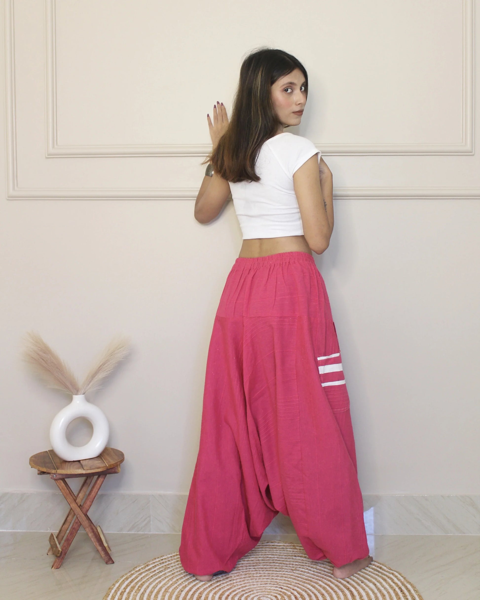 Woman wearing a white top and pink harem pants standing in a room with a decorative stool and vase.