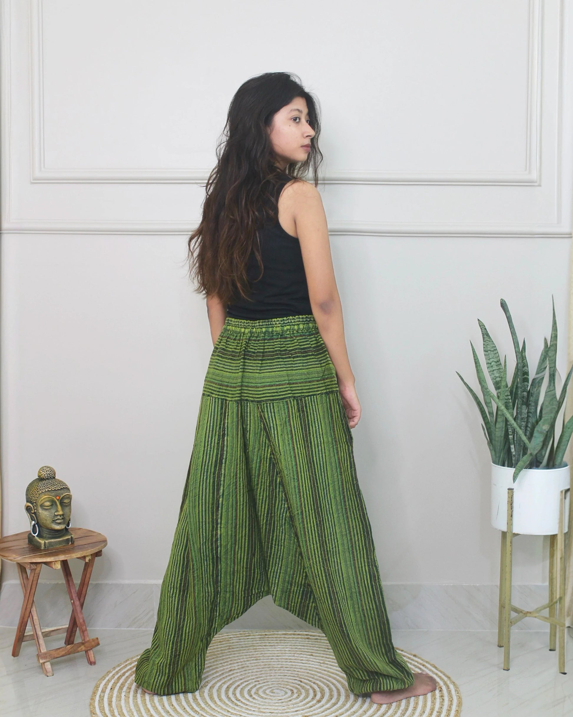 Woman wearing green striped harem pants and a black top in a room with a white wall and small table.