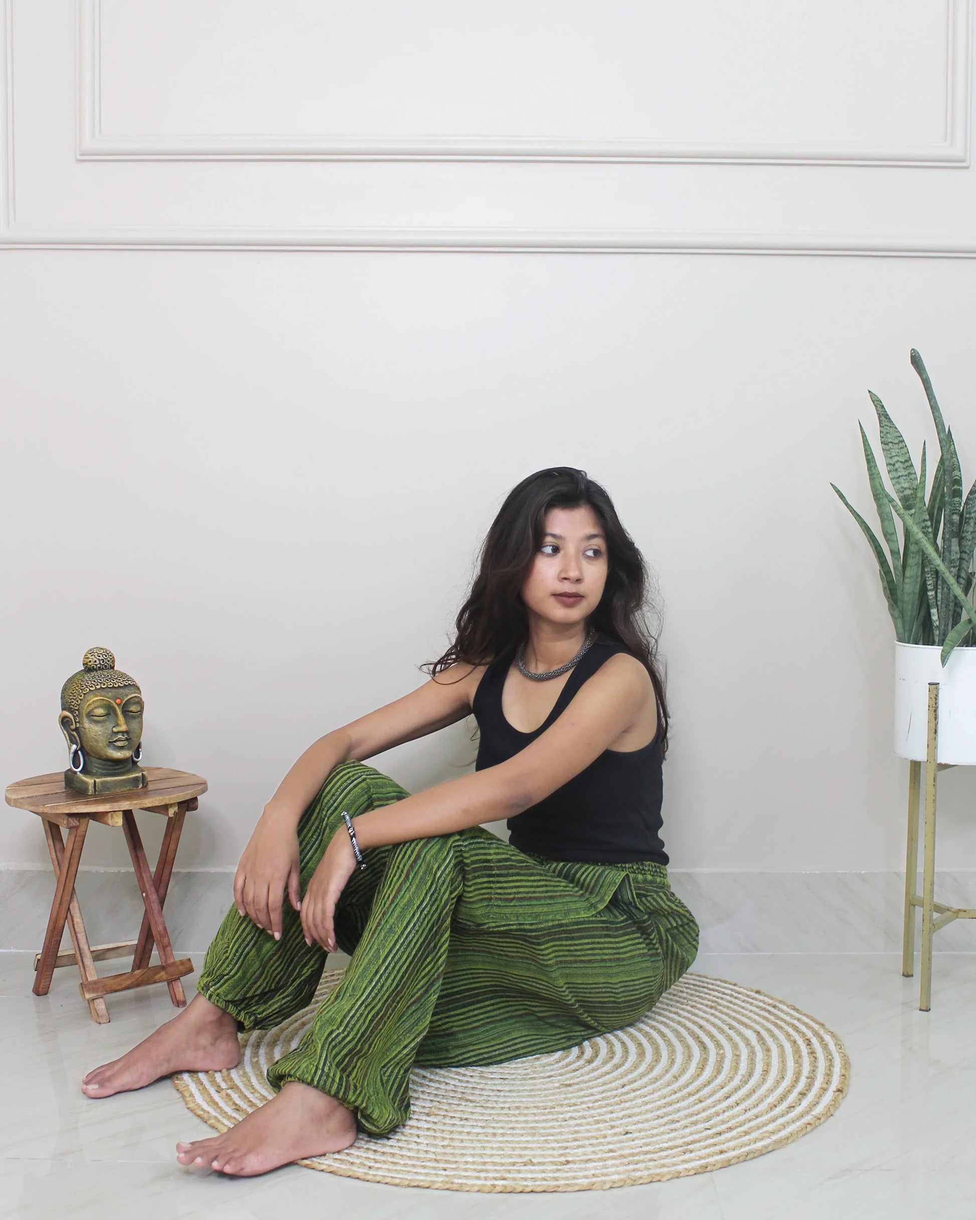 Woman sitting wearing a harem pants on a rug in a room with a plant and small table.
