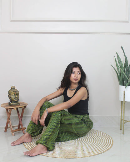 Woman sitting wearing a harem pants on a rug in a room with a plant and small table.