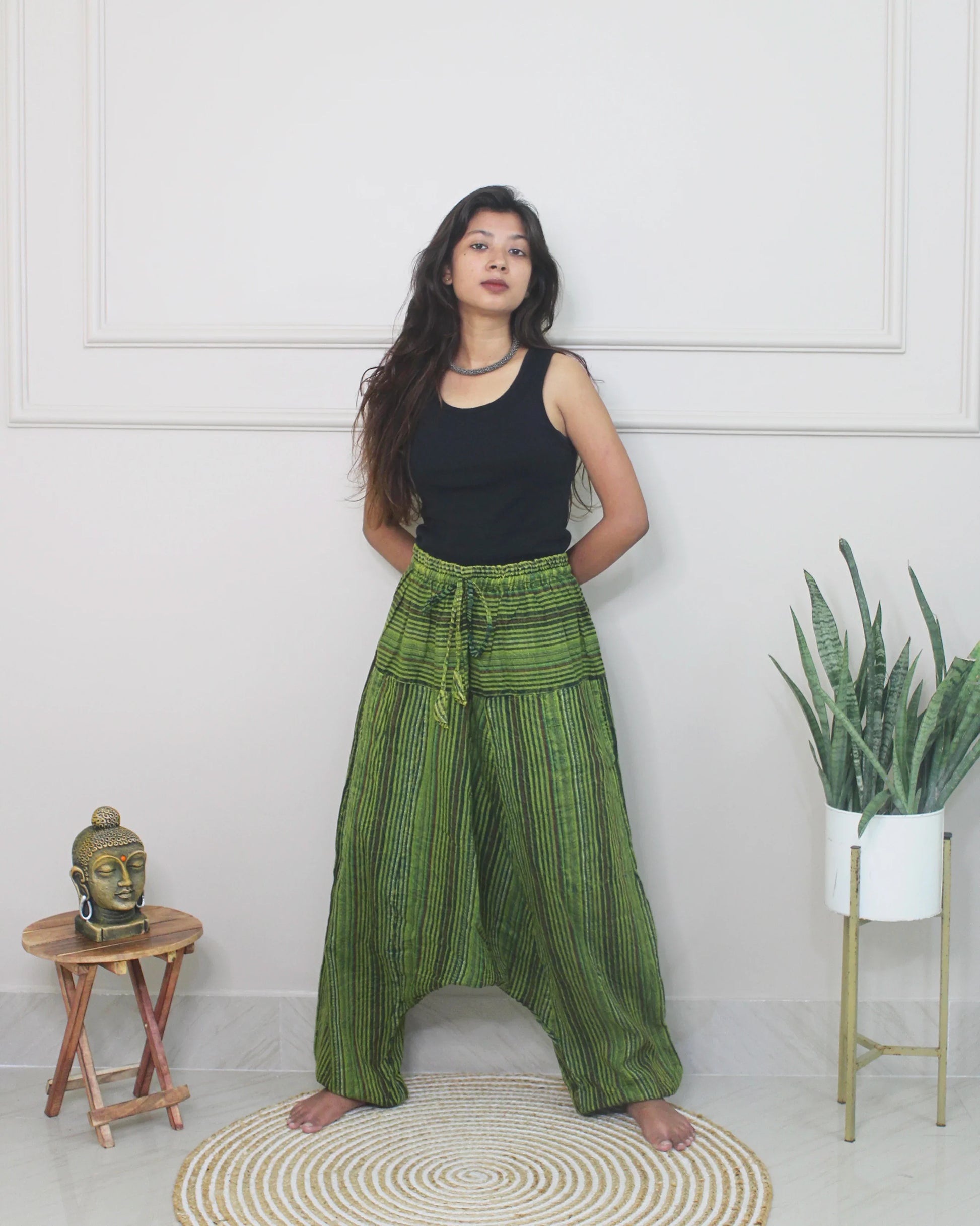 Woman wearing green checkered harem pants and a black top standing in a room with a white wall, small table, and plant.