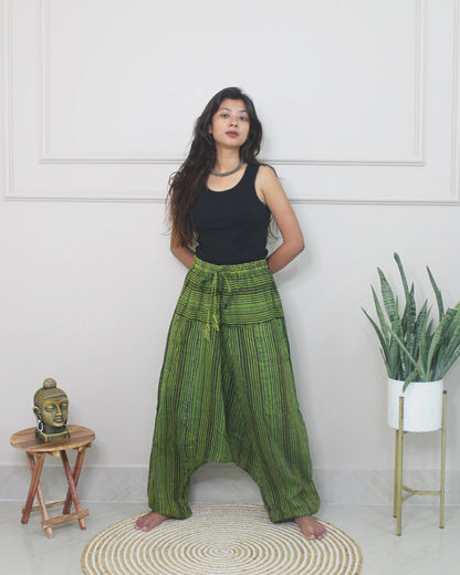 Woman wearing green checkered harem pants and a black top standing in a room with a white wall, small table, and plant.