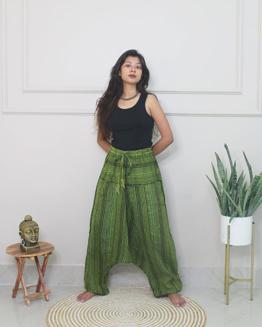 Woman wearing green checkered harem pants and a black top standing in a room with a white wall, small table, and plant.
