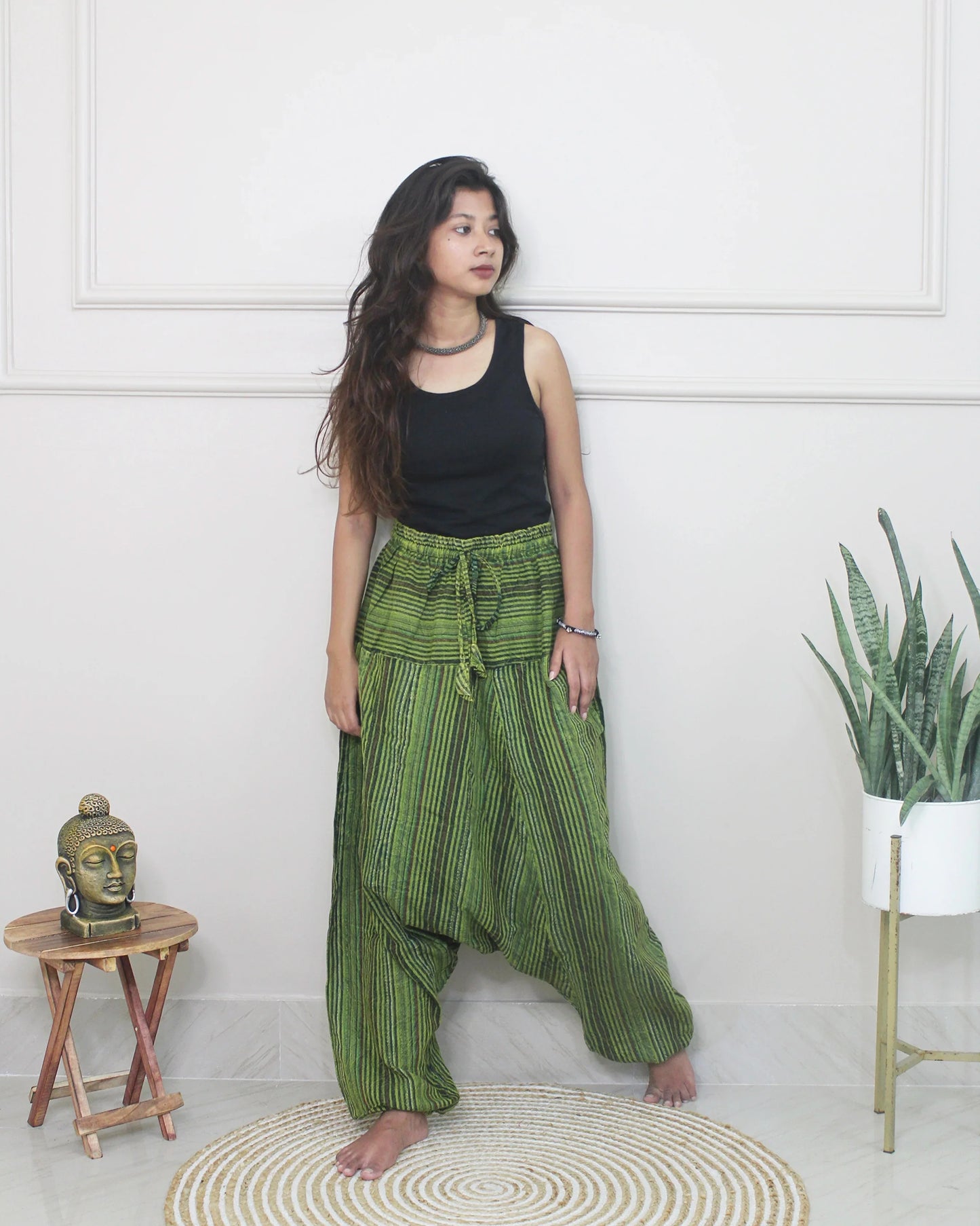 Woman wearing green striped harem pants and a black top standing in a room with a white wall and a small table with a Buddha statue.