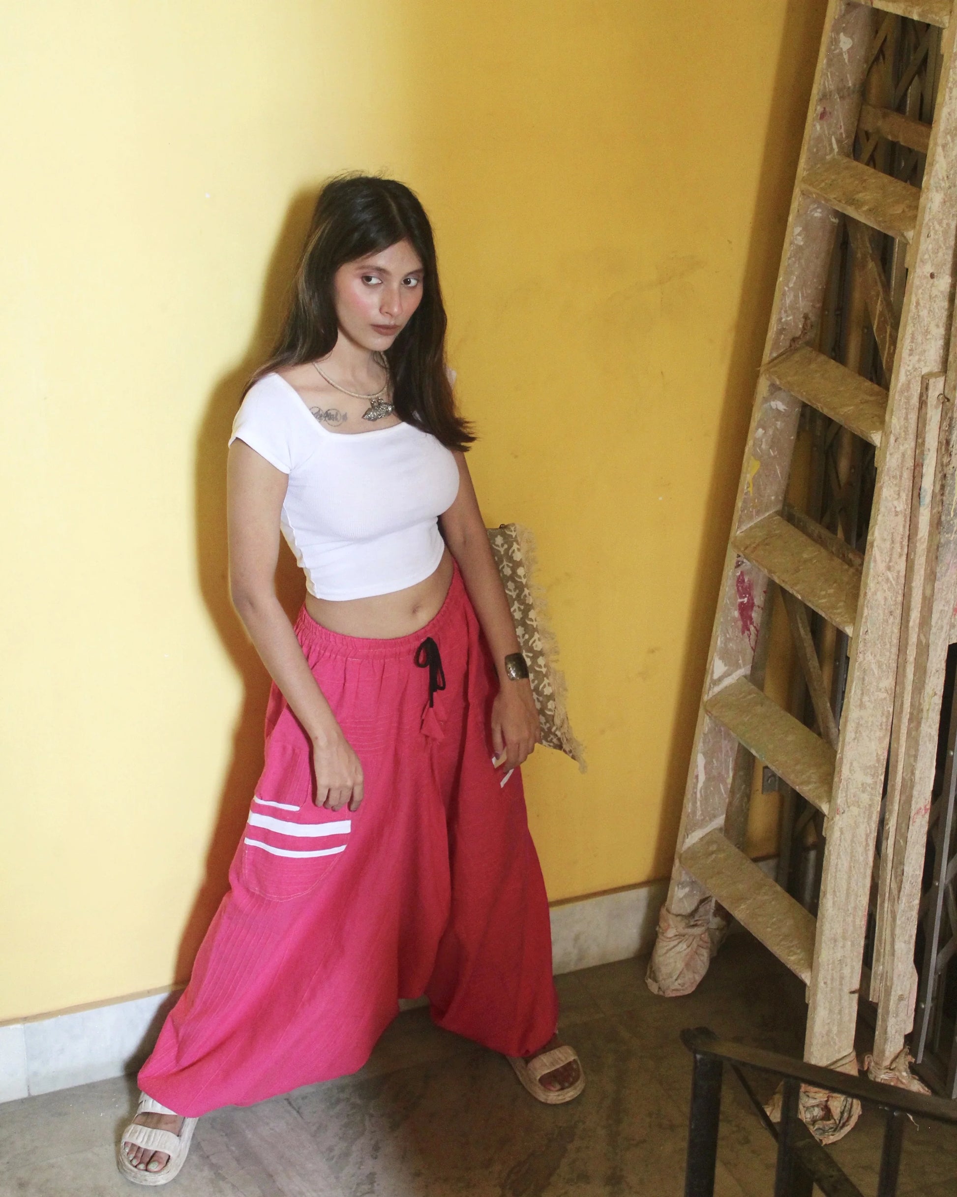 Woman in white top and pink harem pants standing against a yellow wall with a wooden ladder.