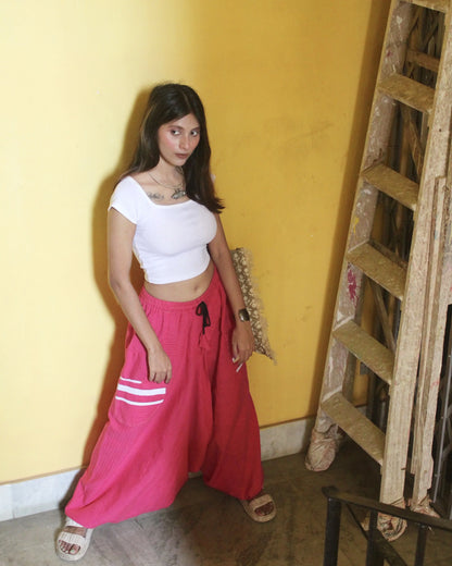 Woman in white top and pink harem pants standing against a yellow wall with a wooden ladder.