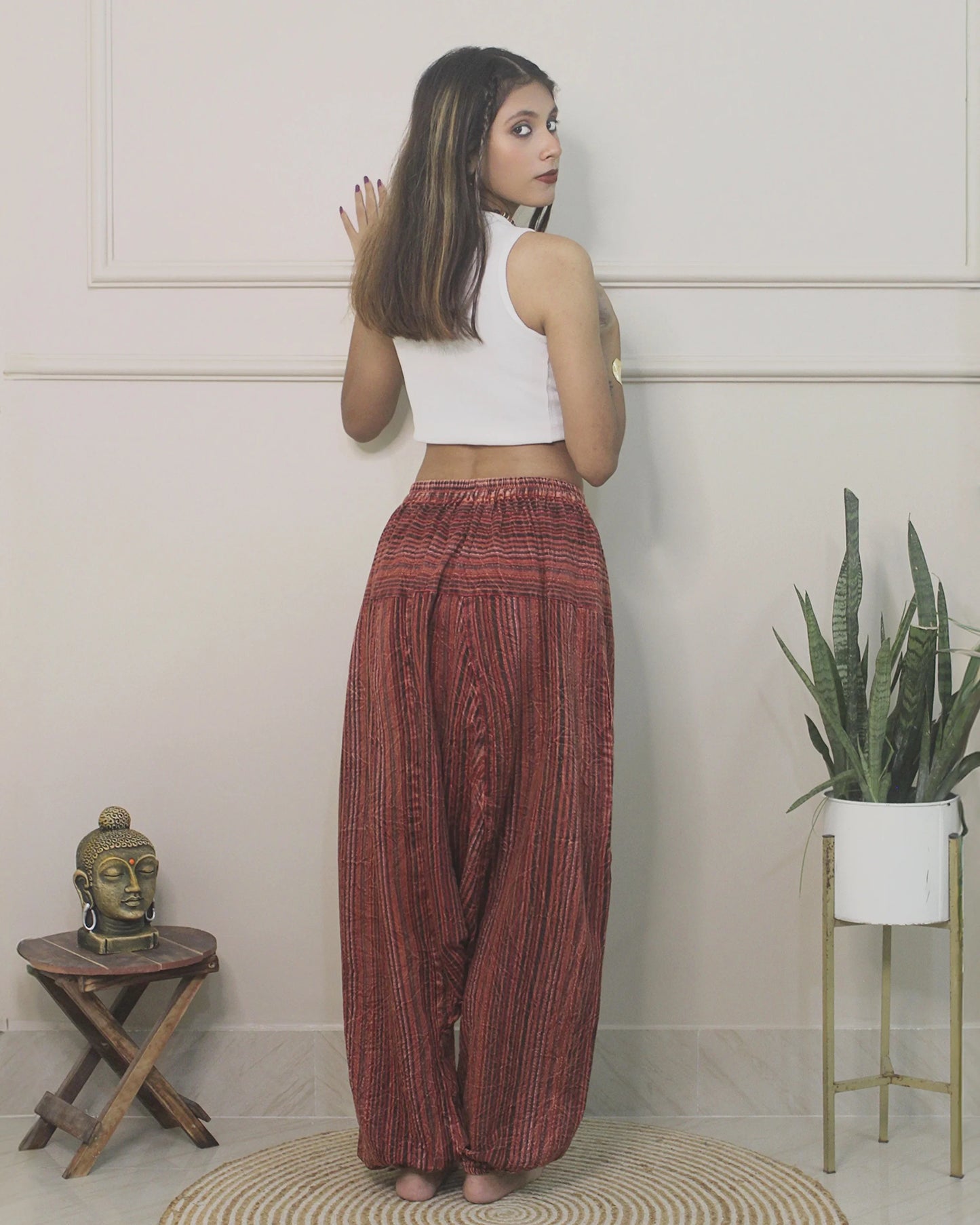 Woman wearing a white top and red  harem pants standing in a room with a plant and small table.