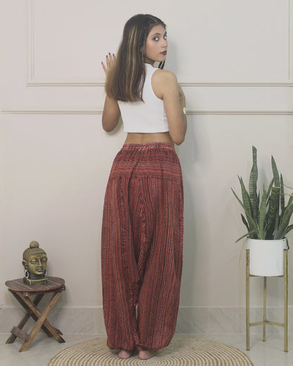 Woman wearing a white top and red  harem pants standing in a room with a plant and small table.