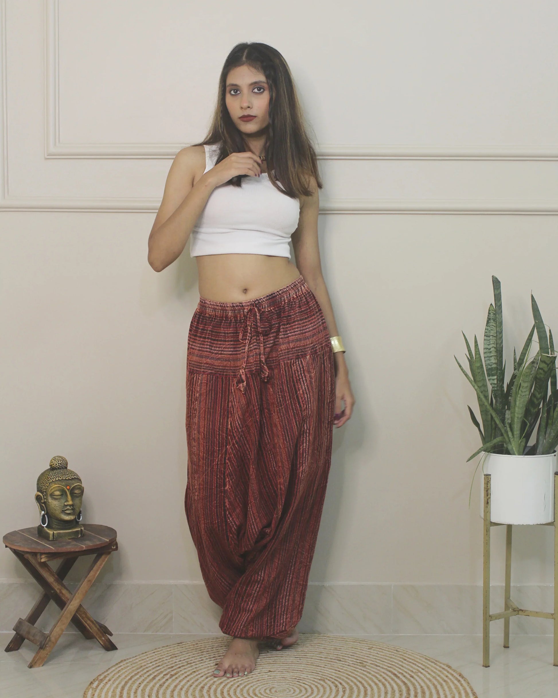 Woman wearing a white top and red harem pants standing in a room with a plant and small table.