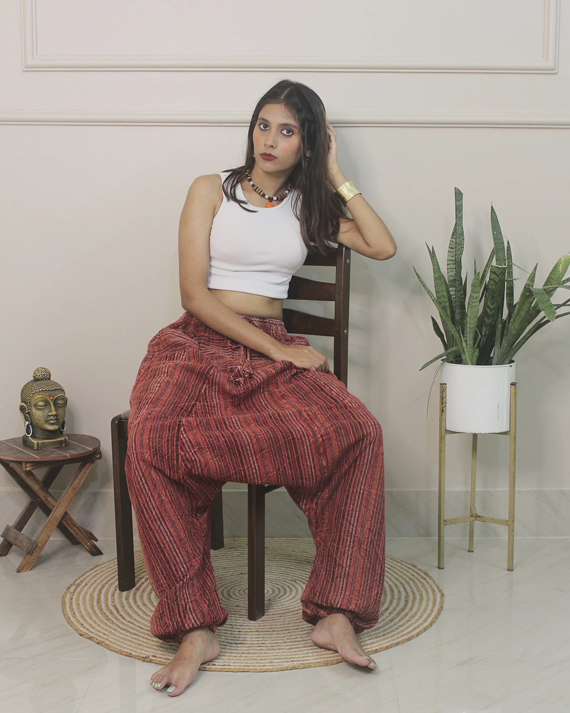 Woman sitting on a chair wearing a white top and harem pants in a room with a plant and small table.