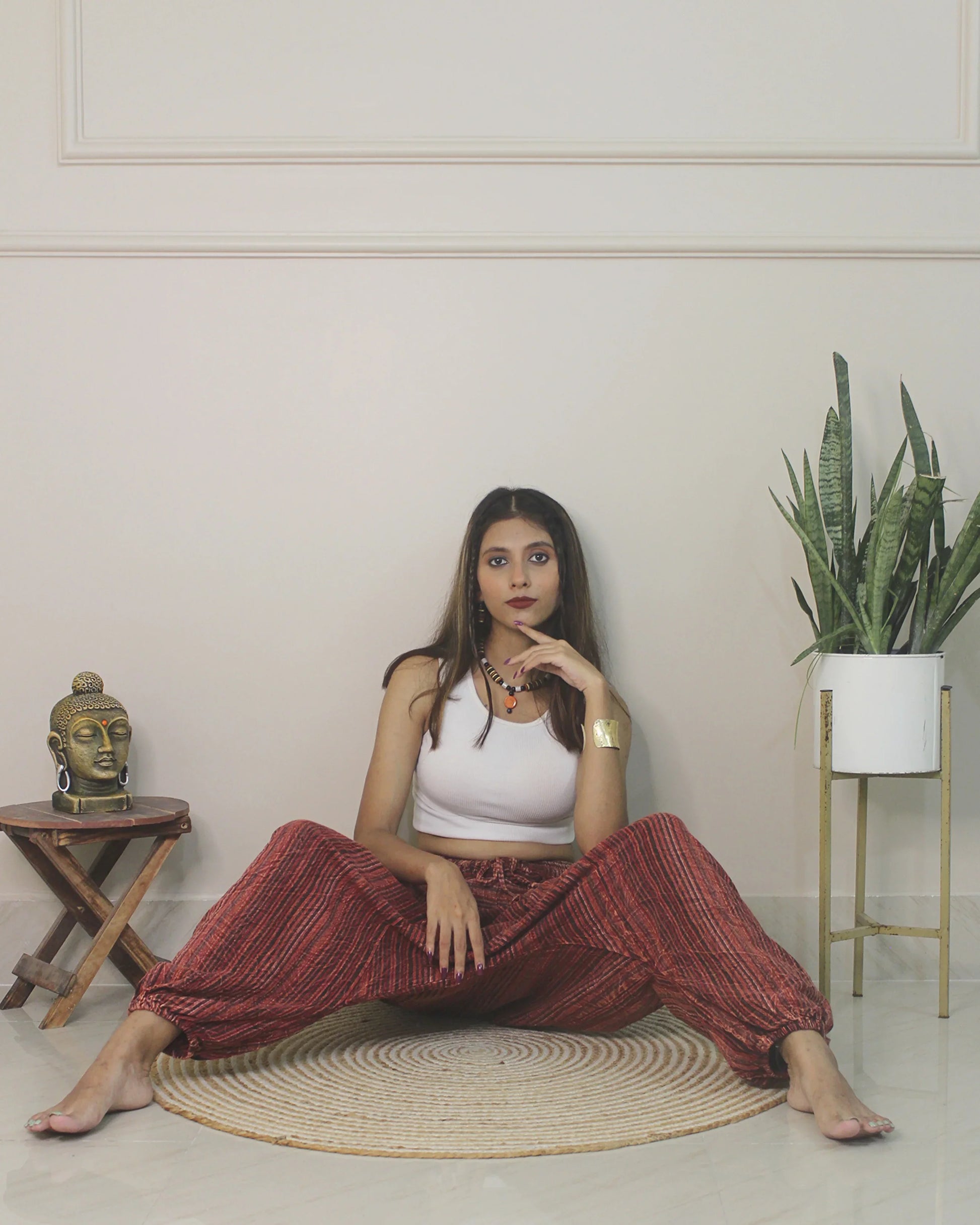 Woman sitting on a woven mat in a room wearing harem pants with a plant and small table.