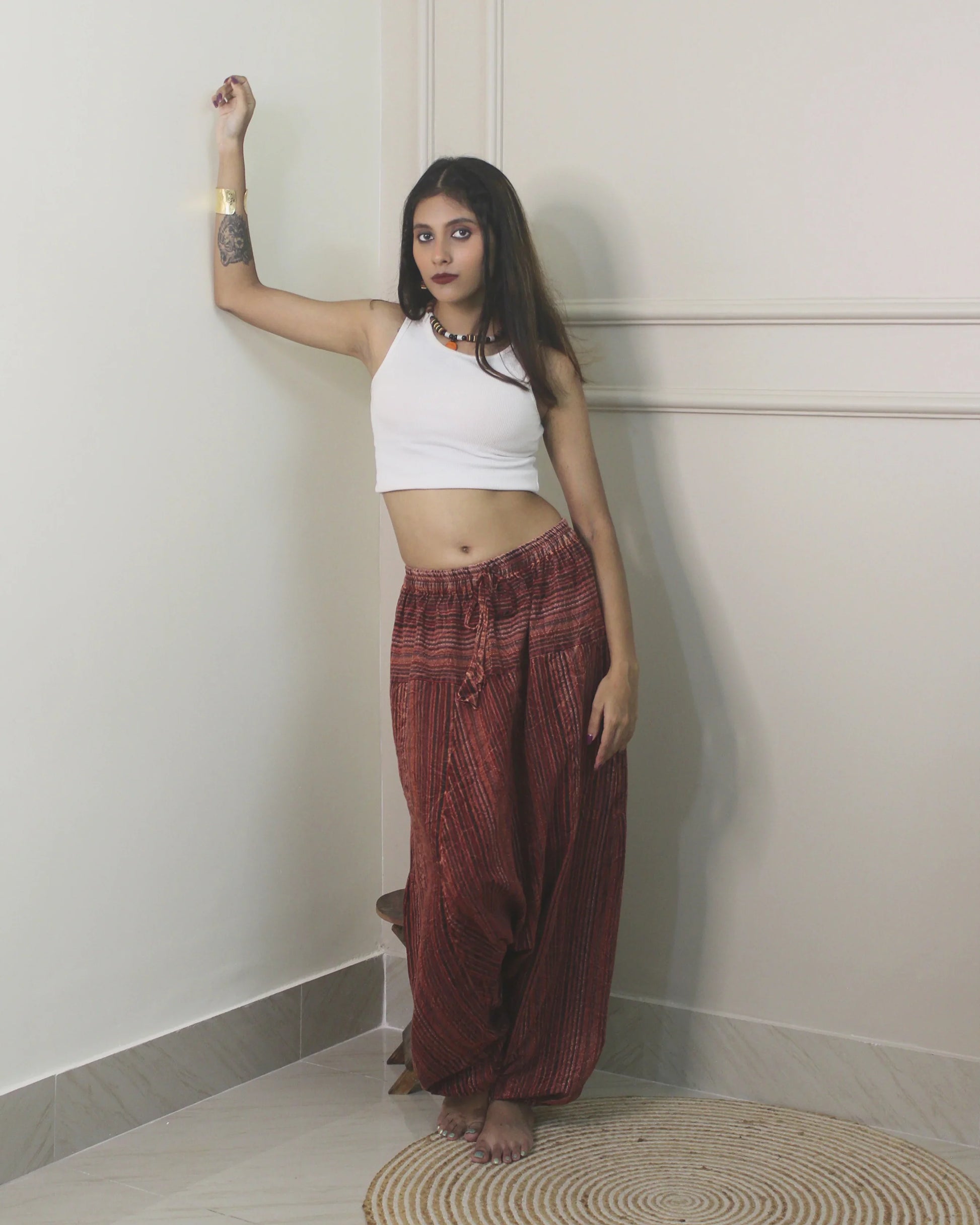 Woman wearing a white top and harem pants standing against a plain wall.