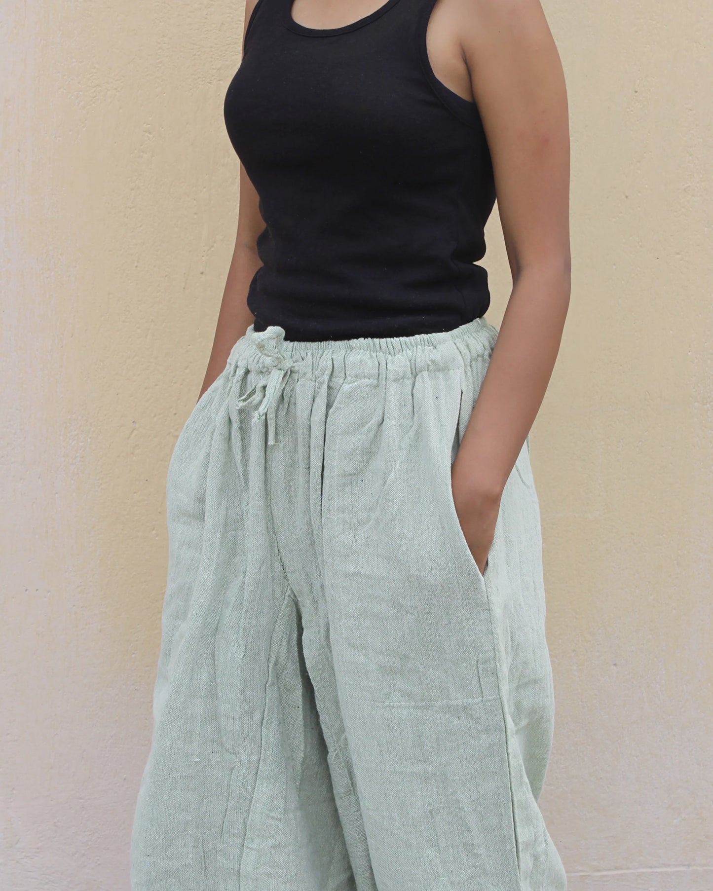 Person wearing a black tank top and light green harem pants against a beige background