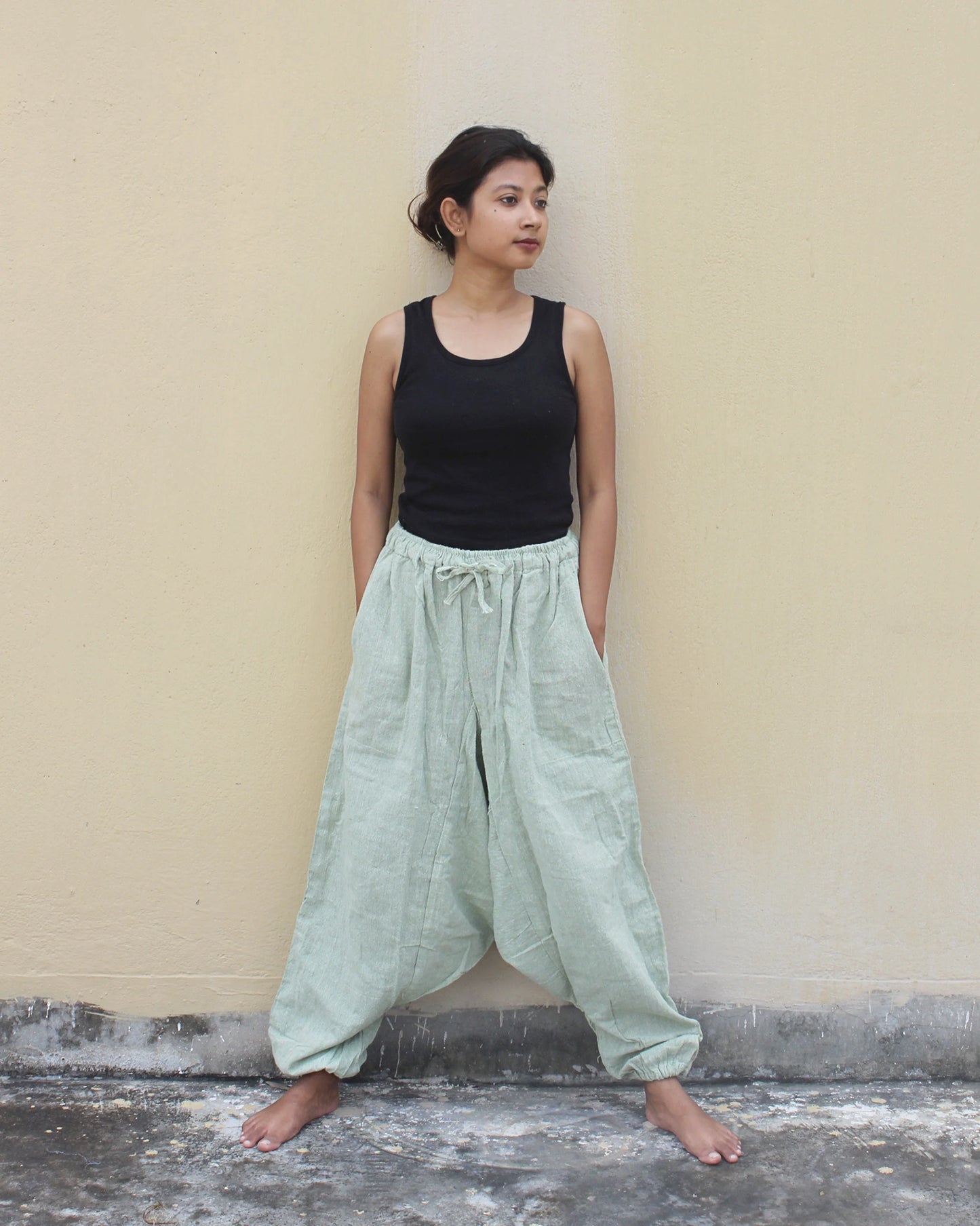 Woman wearing a black tank top and light green wide-leg harem pants against a beige wall.