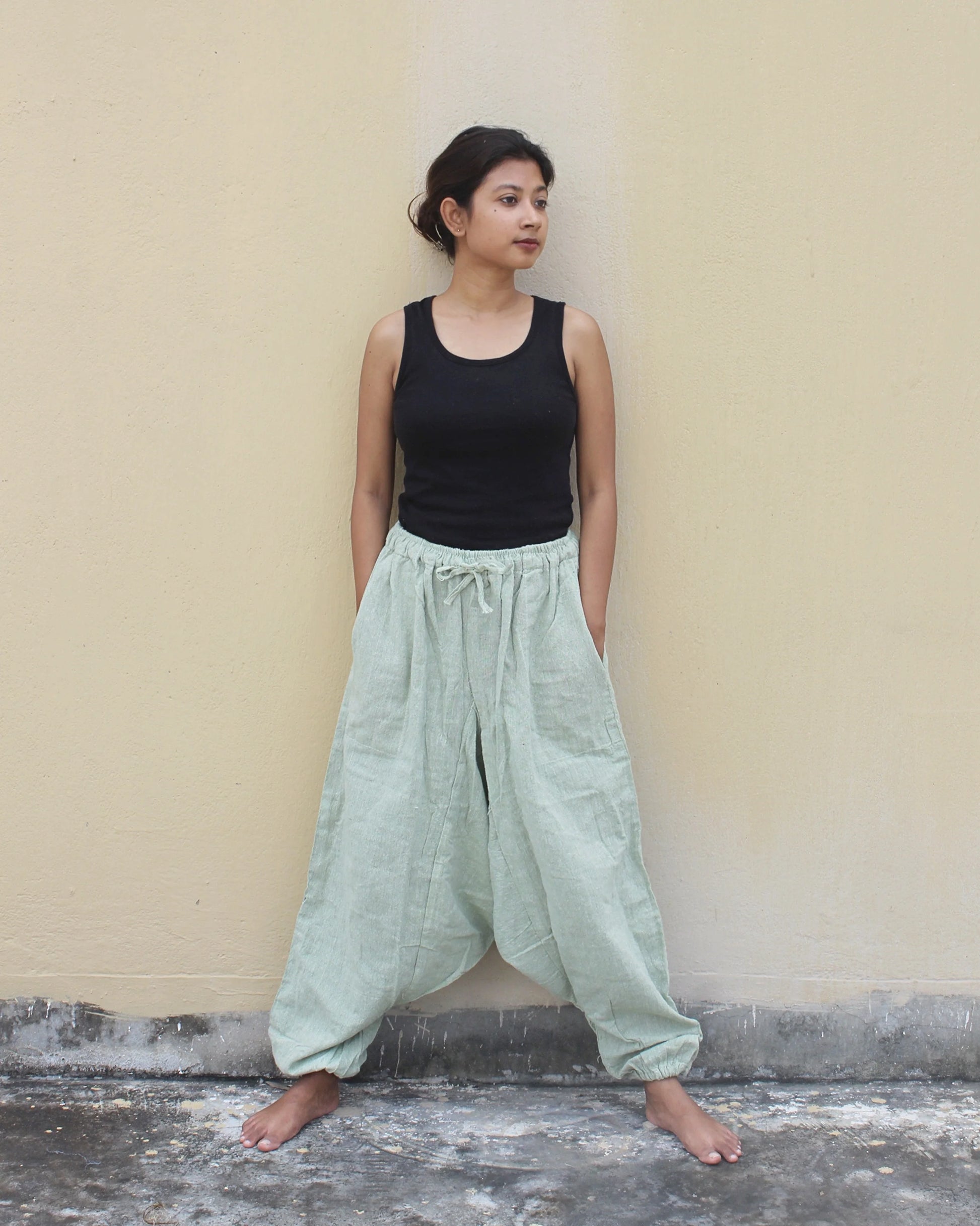 Woman wearing a black tank top and light green wide-leg harem pants against a beige wall.