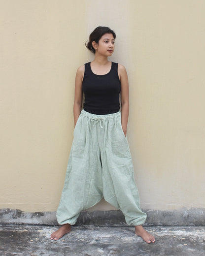 Woman wearing a black tank top and light green wide-leg harem pants against a beige wall.