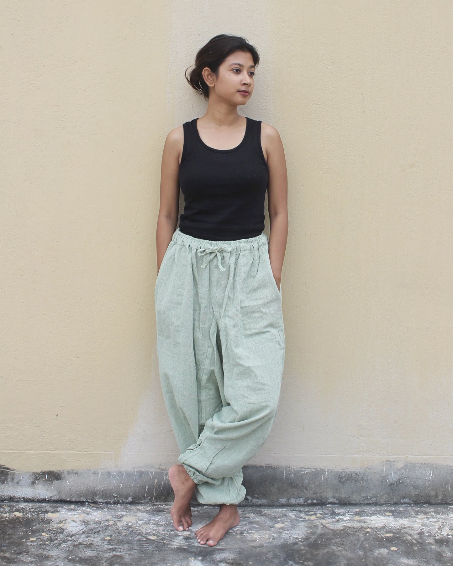 Woman wearing a black tank top and light green harem pants against a beige wall.