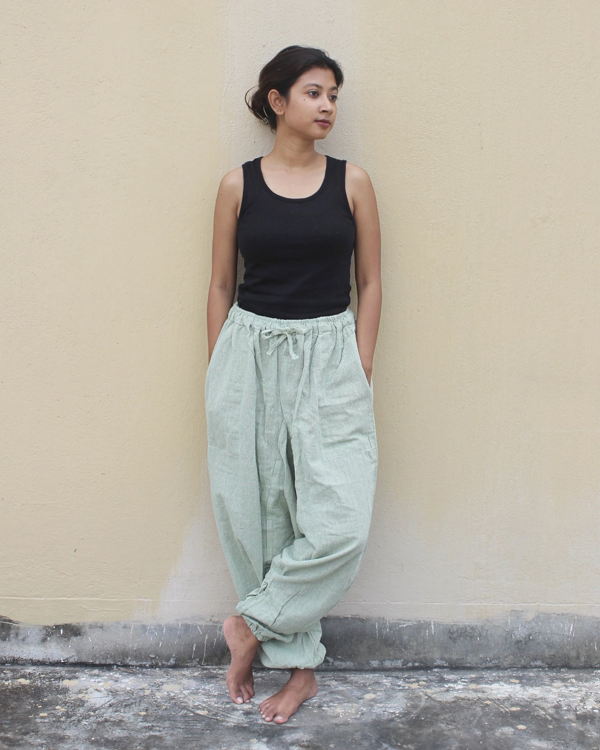 Woman wearing a black tank top and light green harem pants against a beige wall.
