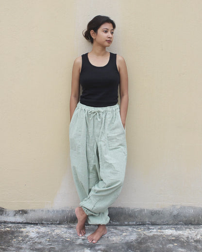 Woman wearing a black tank top and light green harem pants against a beige wall.