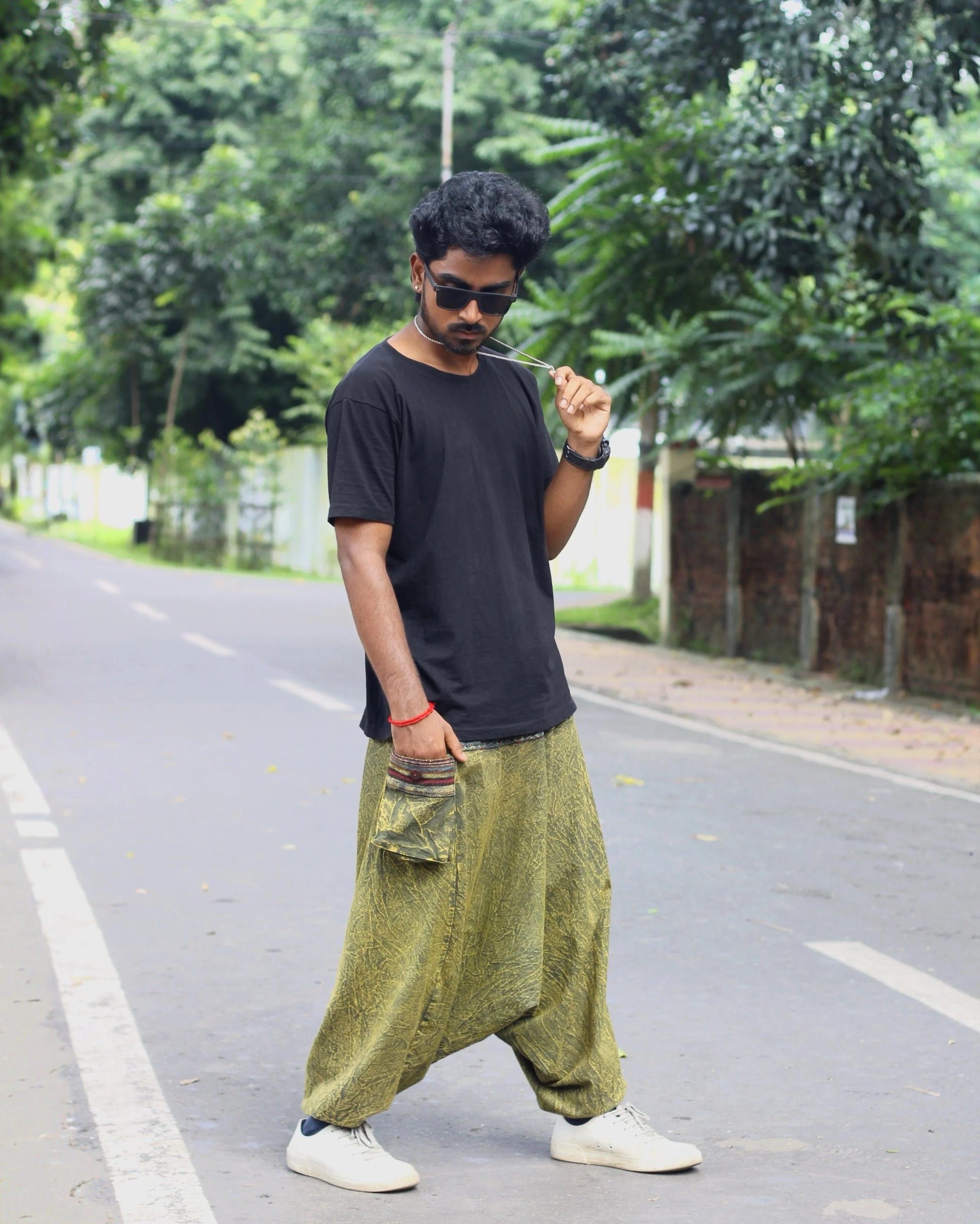 Person wearing a black t-shirt and green baggy harem pants standing on a road with trees in the background