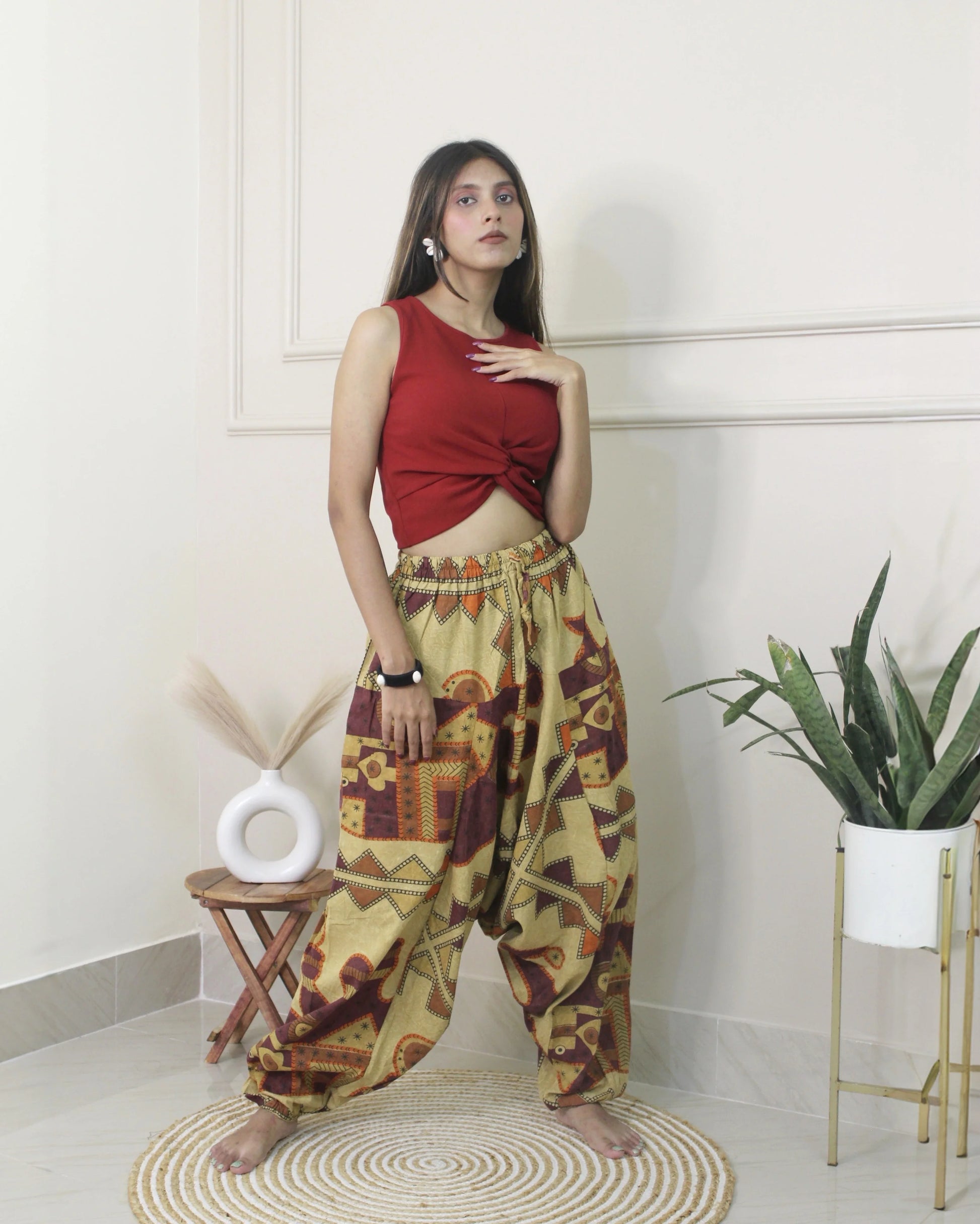 Woman wearing a red top and patterned harem pants standing in a room with a plant and stool.