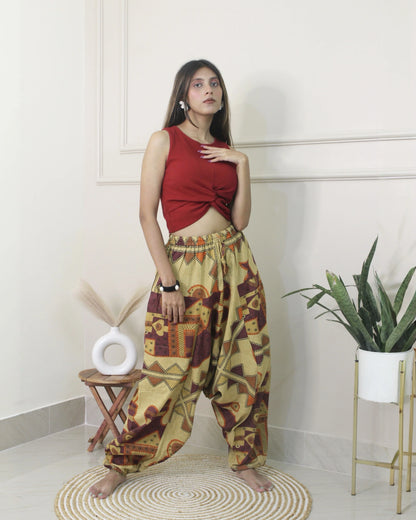 Woman wearing a red top and patterned harem pants standing in a room with a plant and stool.