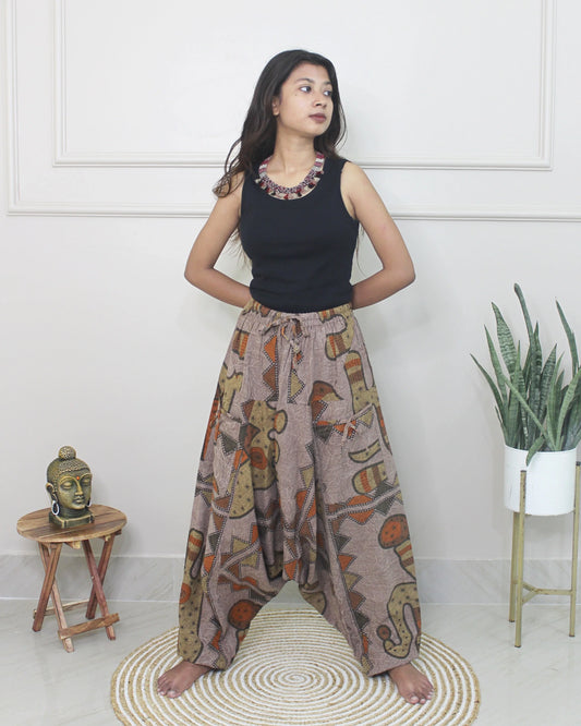Woman wearing a black sleeveless top and patterned harem pants standing in a room with a white wall, small table, and plant.