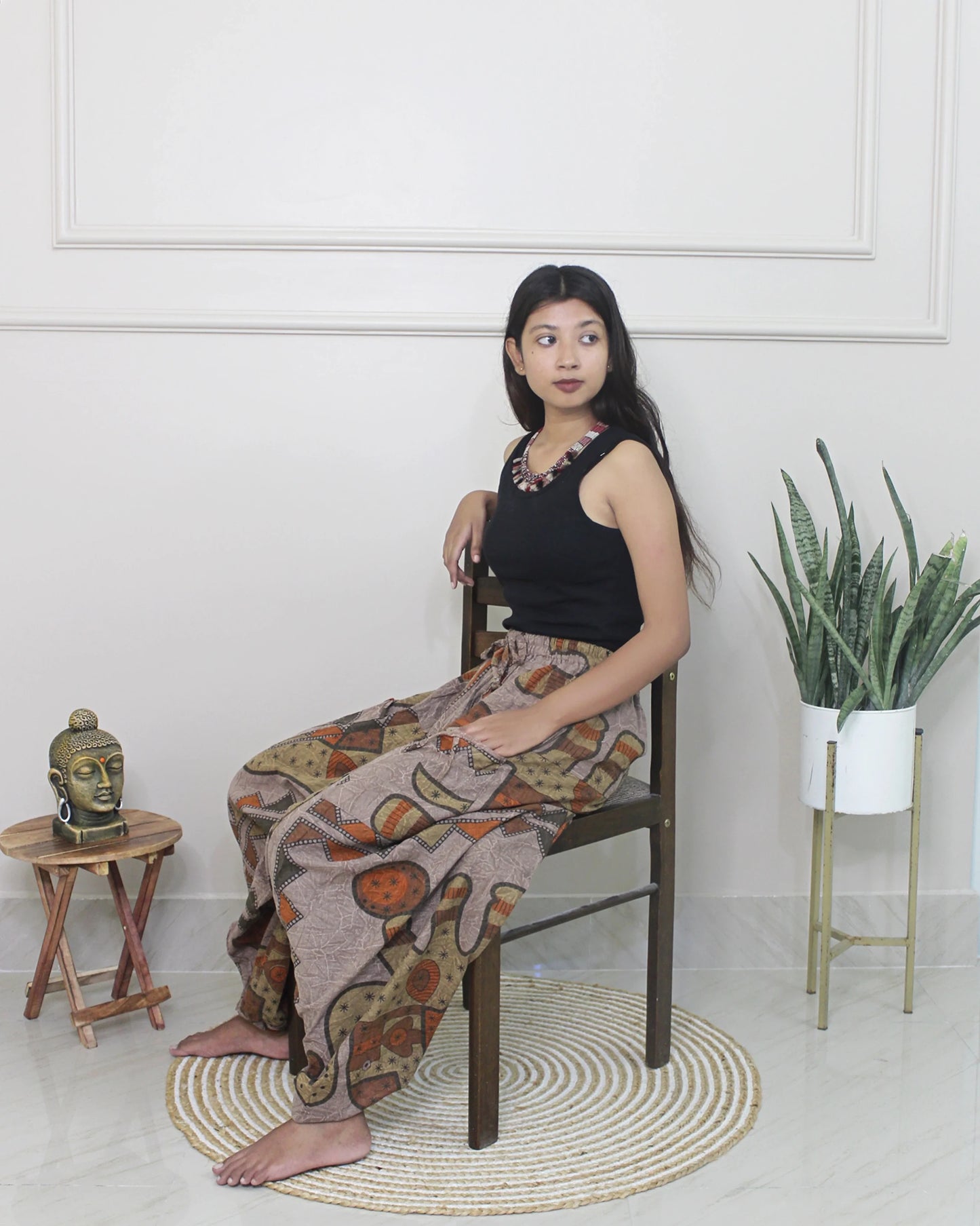 Woman sitting on a chair wearing a black top and patterned harem pants in a minimalistic room.