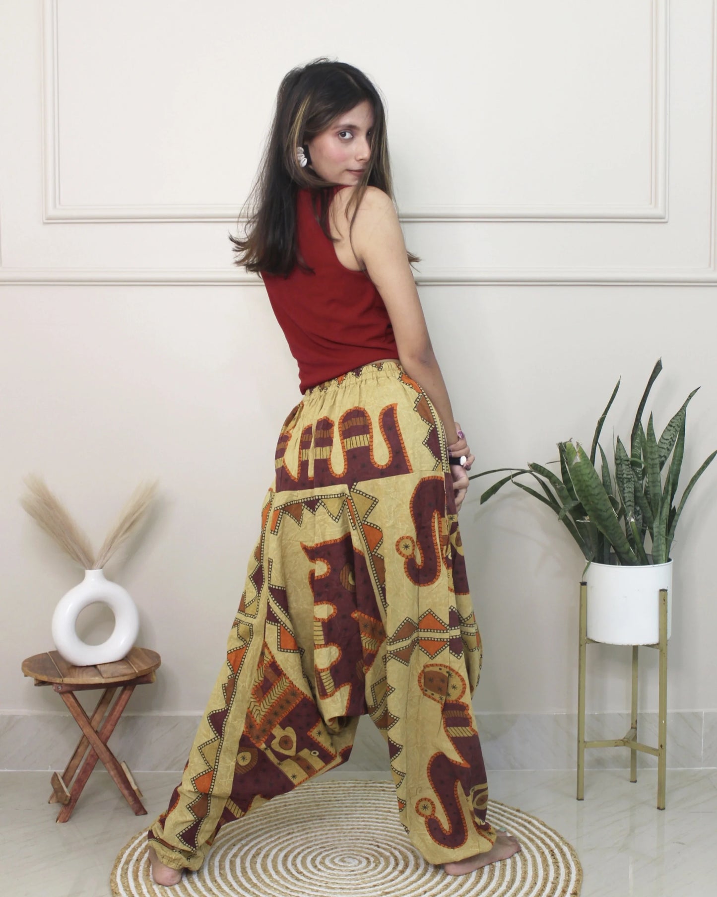 Woman wearing a red top and patterned harem pants in a room with a plant and small table.