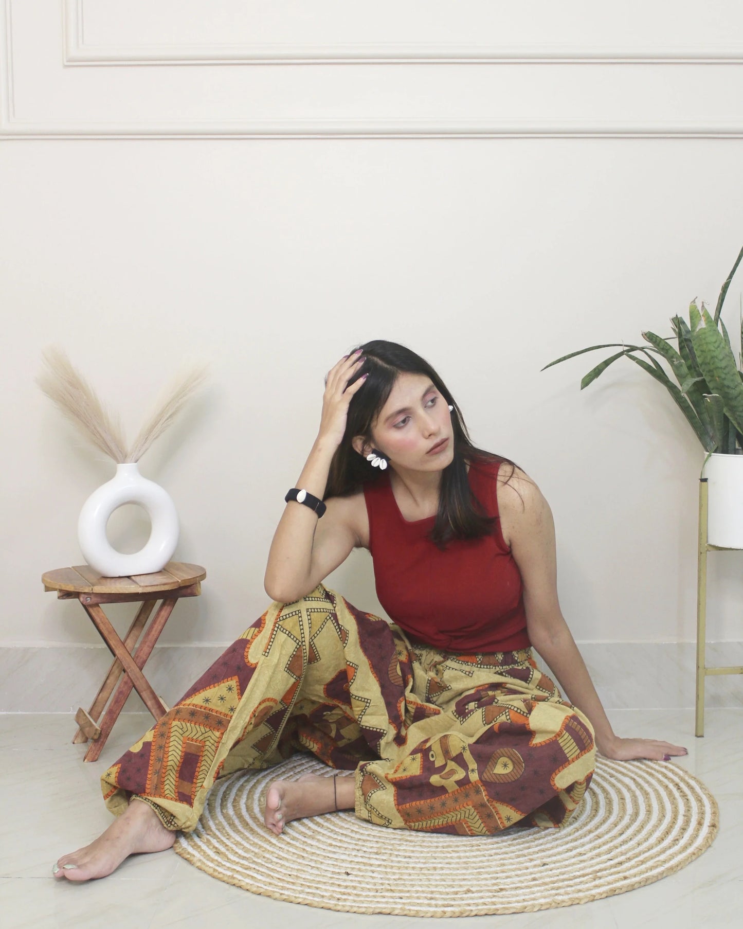 Woman sitting wearing a harem pants on a woven mat in a room with a plant and small table.