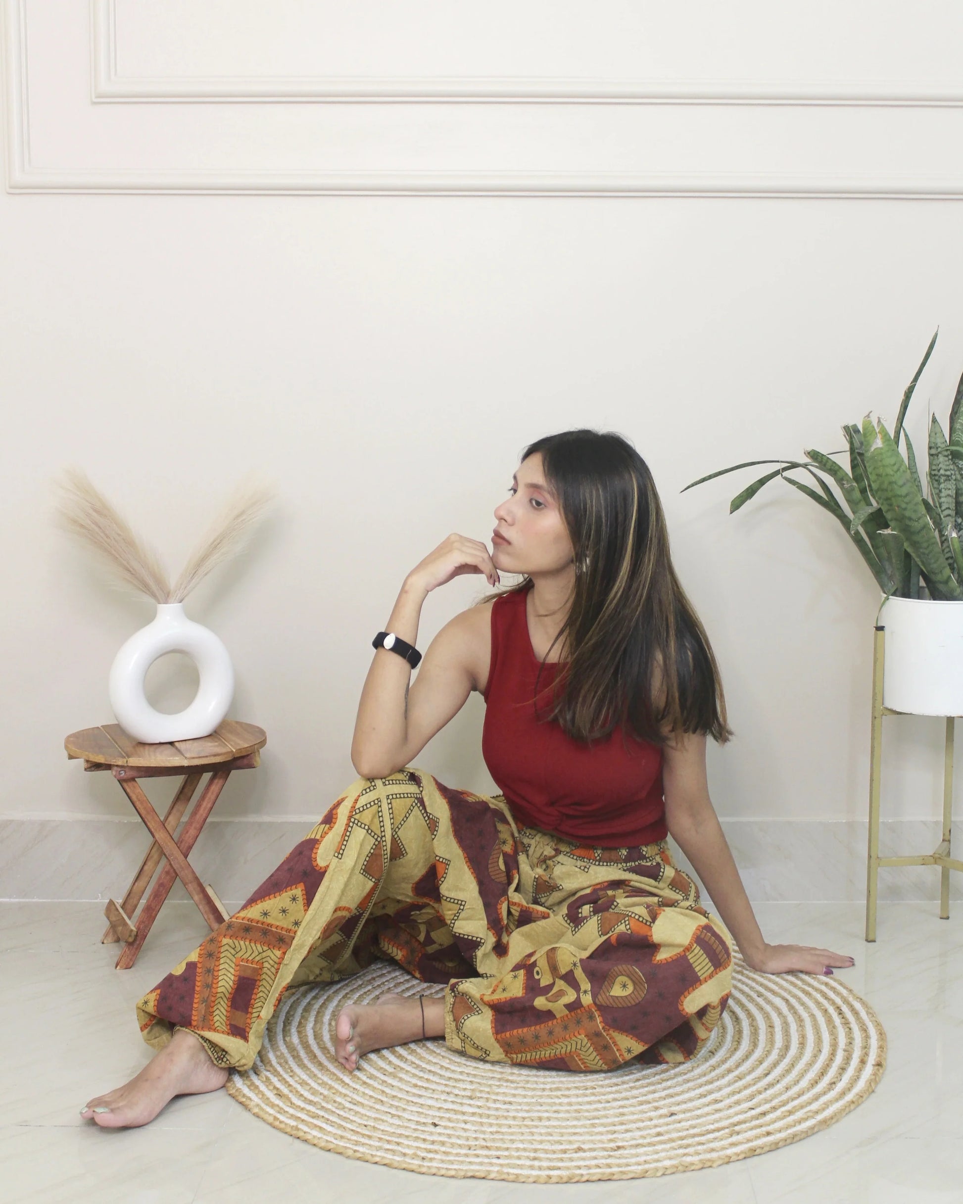 Woman sitting wearing a harem pants on a woven mat in a room with plants and a small table.